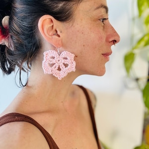 May include: A pair of light pink crochet earrings featuring a six-petaled flower design. The earrings are crafted from yarn and hang from silver-toned metal hooks. The intricate, openwork pattern creates a delicate, lace-like appearance on each earring.