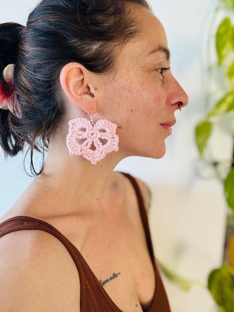 May include: A pair of light pink crochet earrings featuring a six-petaled flower design. The earrings are crafted from yarn and hang from silver-toned metal hooks. The intricate, openwork pattern creates a delicate, lace-like appearance on each earring.
