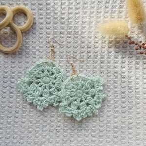 May include: A pair of light mint green crochet earrings featuring a circular, floral-inspired pattern. Each earring is attached to a gold-toned metal hook. The delicate yarn work displays intricate stitching, creating a soft, textured appearance.