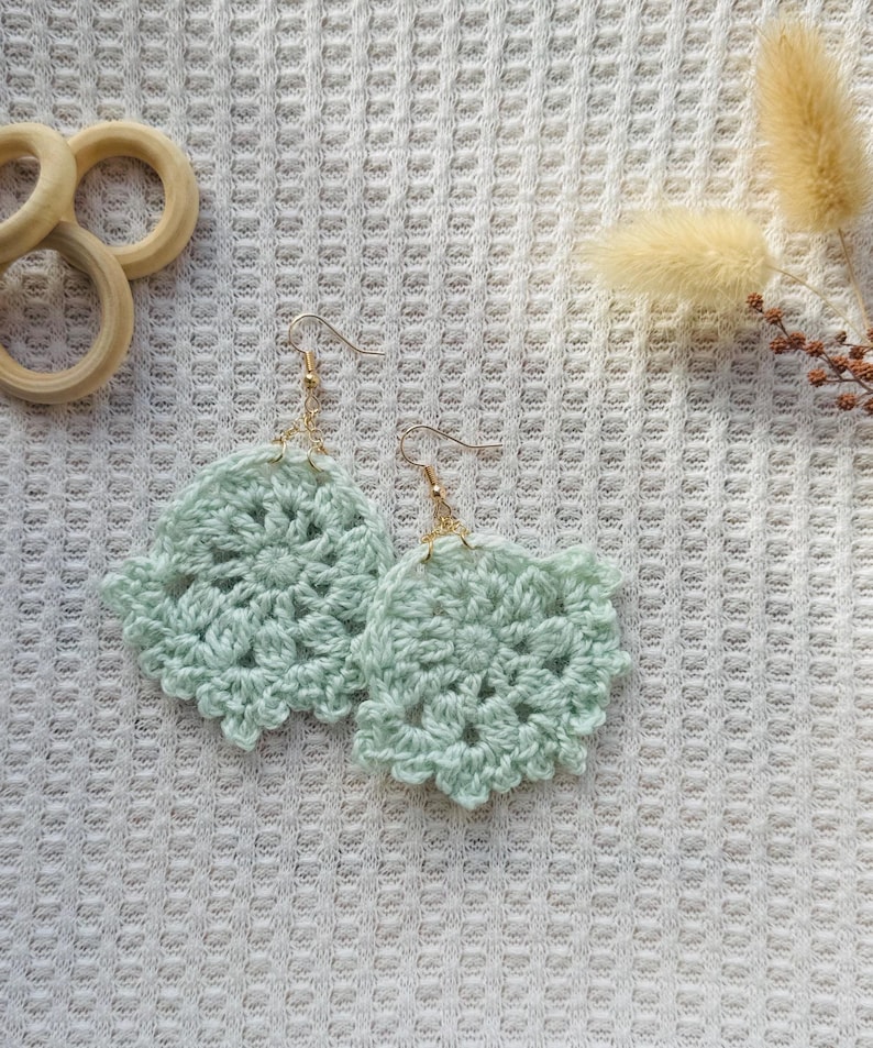 May include: A pair of light mint green crochet earrings featuring a circular, floral-inspired pattern. Each earring is attached to a gold-toned metal hook. The delicate yarn work displays intricate stitching, creating a soft, textured appearance.