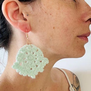 May include: A pale mint green crochet earring featuring a circular, openwork floral pattern. The lightweight, circular textile piece is attached to a thin gold-toned metal hook, designed for insertion into a pierced earlobe for decorative wear.