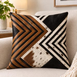 May include: A square throw pillow featuring a geometric pattern made of stitched cowhide panels. The design consists of alternating stripes in black, brown, and white, with some sections displaying natural spotted fur textures.