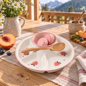 May include: A round, white ceramic divided serving tray with a pink rim and peach illustrations. The tray features a central bowl containing two pink macarons and a wooden spoon resting across the centre. The design includes multiple sections.