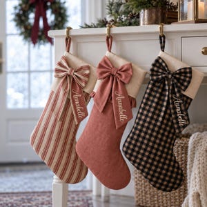May include: Three Christmas stockings hanging from a white mantel. The left stocking has red and beige stripes, the center is solid dusty rose, and the right is black and beige gingham. Each features a large bow with the name Annabelle.