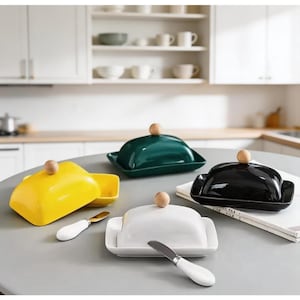 May include: Four rectangular butter dishes with lids in yellow, dark green, black, and white. Each lid features a small, round, light-colored wooden knob on top. Two silver-toned butter knives with white handles are placed next to the dishes.