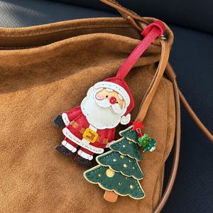 Könnte beinhalten: Eine braune Wildledertasche mit einem hängenden Anhänger, bestehend aus einer rot-weißen Weihnachtsmannfigur aus Leder und einem dunkelgrünen Weihnachtsbaum aus Leder mit goldenen Akzenten, einem kleinen roten Stern und grünen Glasperlen.