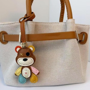 Può includere: Una borsa in tela beige con manici in pelle marrone e una cinghia orizzontale con finiture color argento. Un ciondolo a forma di orsetto multicolore pende dalla cinghia, con testa marrone, muso bianco e arti gialli, blu e rosa.