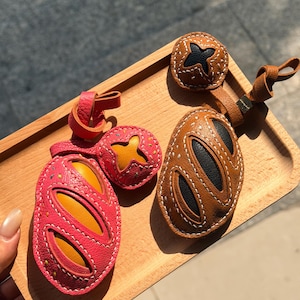 May include: Two leather keychains shaped like loaves of bread, one in bright pink with yellow accents and one in brown with black accents. Each keychain features a small circular charm with a star-shaped cutout and a leather loop for attachment.
