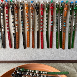 Pode incluir: Uma coleção de tiras de couro em várias cores, incluindo preto, castanho, vermelho e verde, cada uma com fivelas de metal e tachas decorativas prateadas, detalhes em forma de estrela ou incrustações circulares turquesa.