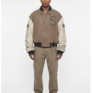May include: A brown varsity jacket with off-white sleeves, a black collar, and a large letter S patch on the chest with hanging threads. The jacket features the number 2024 on the sleeves. It is paired with matching brown cargo trousers and brown trainers.