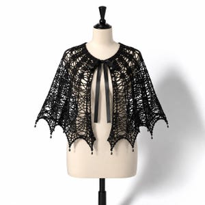 May include: A black crochet capelet featuring an intricate web-like pattern with a scalloped hemline. The garment is fastened at the neckline with a thin black satin ribbon tie and is finished with small, dangling black teardrop-shaped beads.