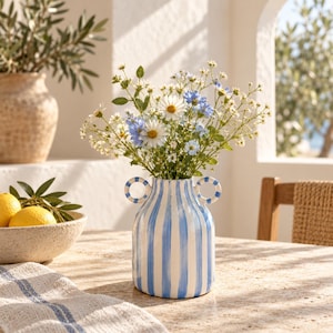 May include: A ceramic vase featuring vertical blue and white stripes with two small circular handles on the sides. The vase is filled with a bouquet of white daisies and small blue flowers, creating a bright and colorful floral arrangement.