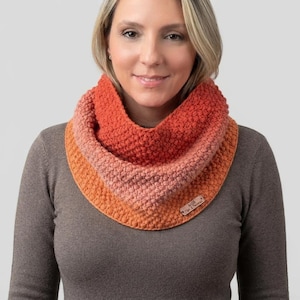May include: A textured infinity scarf featuring a gradient of orange and pink hues. The knit fabric displays a consistent bumpy pattern throughout. A small rectangular brown leather tag is attached to the lower right side with the text "A&M Time & Thread".