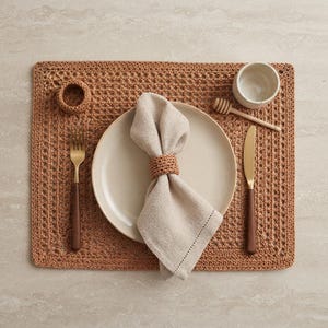 May include: A rectangular brown crocheted placemat featuring a central beige ceramic plate, a linen napkin held by a matching crocheted ring, a gold-toned fork and knife with dark wooden handles, a small ceramic bowl, and a wooden honey dipper.
