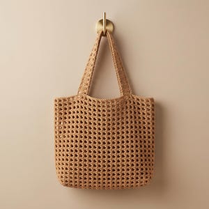 May include: A tan crochet tote bag featuring an open-weave grid pattern and two integrated shoulder straps. The bag is constructed from a textured yarn material and displays a consistent square-holed design throughout the body and handles.