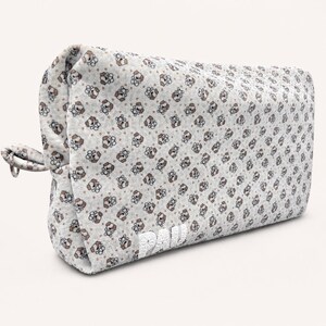 Peut inclure: Une trousse rectangulaire à fermeture éclair avec un motif répétitif de petits visages de chiots bruns et blancs et d'empreintes de pattes sur un fond gris clair. Le mot PAU est brodé en fil blanc sur le coin inférieur avant de l'accessoire.