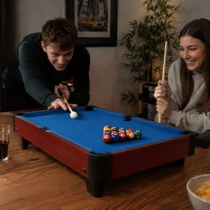 May include: A tabletop pool table with a blue felt surface, dark wood-toned frame, and black corner accents. The set includes a white cue ball, a wooden cue stick, and a triangular rack of multicolored numbered billiard balls arranged on the table.