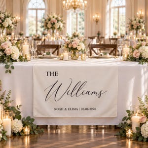 Puede incluir: Un camino de mesa de tela blanca con el texto impreso en negro THE Williams, seguido de NOAH & ELISIA | 06.06.2026. El camino está colocado sobre una mesa decorada con centros de mesa florales, portavelas de cristal y velas blancas.