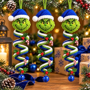 May include: Three identical crochet hanging ornaments featuring a green character head wearing a blue and white Santa hat. The body is a long, spiraling tube in blue, green, and white, decorated with small holly leaves, red berries, and candy canes.