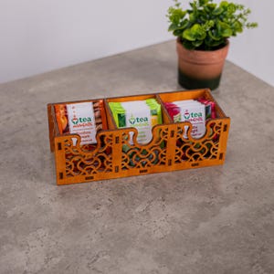 Puede incluir: Un organizador rectangular de madera para bolsitas de té con tres compartimentos y un intrincado patrón geométrico cortado con láser en el panel frontal. El organizador contiene varias bolsitas de té Tea Moments en envases de color naranja, verde y rojo.