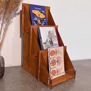 May include: A tiered wooden display stand with three levels, featuring a dark brown finish. The stand holds three rectangular cards, with the top card displaying the word Ukraine. A clear acrylic front panel is attached to the lowest tier.