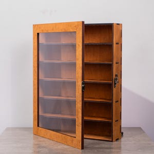 May include: A rectangular wooden display case with a light brown burl wood finish. The case features a hinged door with a clear glass panel, a metal latch, and five internal shelves for storage. The side panels show dark brown joinery details.