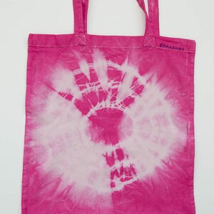 Könnte beinhalten: Rosa-farbene Batik-Tragetasche mit einem weißen Handabdruck-Design und dem Text "ShakeMBO" in Lila.