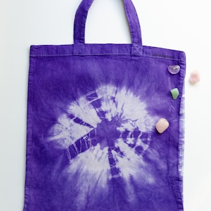 Könnte beinhalten: Lilafarbene Batik-Tragetasche mit weißem Wirbelmuster und dem aufgedruckten Text "SHAKEH8" an der Seite. Die Tasche liegt auf einer weißen Oberfläche mit kleinen rosa und grünen Steinen.