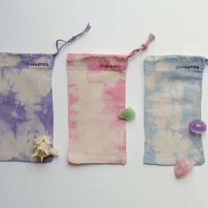 Könnte beinhalten: Drei kleine Stoffbeutel mit Kordelzug. Die Beutel sind in den Farben Lila, Rosa und Blau im Tie-Dye-Verfahren gefärbt. Jeder Beutel trägt den Schriftzug "ShakeMBO". Die Beutel liegen auf einer weißen Oberfläche.