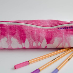 Könnte beinhalten: Rosa-farbene Batik-Federmappe mit einem weißen Reißverschluss und dem in Lila gestickten Wort "Snack Time" auf der Vorderseite. Drei orangefarbene Stifte mit violetten Spitzen liegen vor der Federmappe.