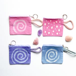Könnte beinhalten: Vier kleine Reißverschlusstaschen mit verschiedenen Batik-Mustern. Die Taschen sind rosa mit weißen Punkten, rosa mit einer weißen Spirale, lila mit einer weißen Spirale und blau mit einer weißen Spirale. Jede Tasche hat einen silbernen Schlüsselring.