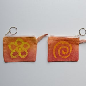 Könnte beinhalten: Zwei kleine orangefarbene Stoffbeutel mit Reißverschluss und Schlüsselringen. Ein Beutel hat ein gelbes Blumendesign und der andere ein gelbes Spiralendesign.