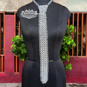 Peut inclure: Une cravate argentée fabriquée à partir d'anneaux métalliques entrelacés dans un motif de cotte de mailles. La cravate présente un nœud en haut et pend verticalement. Un logo avec le texte CHAINMAIL KINGDOM est visible en haut à gauche.