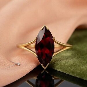 May include: A gold ring featuring a large, deep red marquise-cut gemstone set in a prong setting. The band of the ring splits into two thin, smooth gold arms that meet at the base of the stone, creating a sleek and refined appearance.
