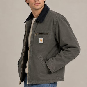 Può includere: Una giacca in tela grigio antracite con colletto in velluto a coste blu scuro, cerniera frontale color ottone e tasca sul petto con chiusura a zip. Una toppa rettangolare del marchio Carhartt è cucita sul lato sinistro del petto.
