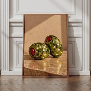 May include: A framed wall art print featuring two oval-shaped objects with a green, pixelated, mosaic-like surface pattern. Each object has a bright, circular red centre. The objects are positioned on a light-coloured, textured surface.