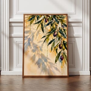 May include: A framed watercolour painting featuring a branch with dark purple and green olives and elongated green leaves. The background is a soft yellow wash with a blue-toned shadow of the olive branch cast across the lower left side.