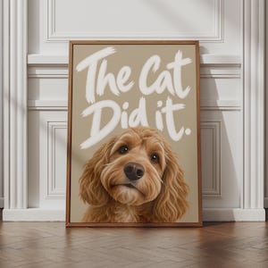 May include: A rectangular framed wall art piece featuring a light brown background with the white text The Cat Did it. centred above a portrait of a golden-brown curly-haired dog with large dark eyes and a black nose looking directly forward.