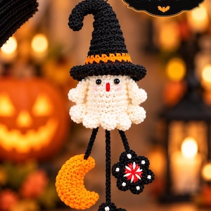 Op de afbeelding: Een gehaakt spookornament met een zwarte heksenhoed met een oranje band. Het spook heeft een wit lichaam, zwarte ogen en een rode mond. Drie zwarte koordjes hangen aan de onderkant, met een oranje halve maan en twee zwarte bloemen.