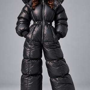 Op de afbeelding: Een zwarte, volledig gevoerde, isolerende gewatteerde jumpsuit met een hoge, oversized kraag, een centrale zilveren ritssluiting en een getailleerde taille met drukknopen. Het kledingstuk heeft lange mouwen met geribbelde boorden.