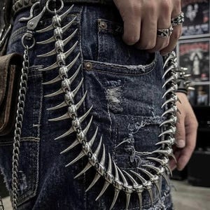 May include: A silver-toned metal wallet chain featuring a series of interconnected, curved, sharp-pointed spikes resembling a spinal column. The chain is attached to a dark blue distressed denim garment with a metal clasp and a secondary thin chain.