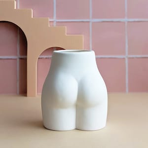 May include: White ceramic planter shaped like a human bottom. The planter is sitting on a light brown surface in front of a pink tiled wall.