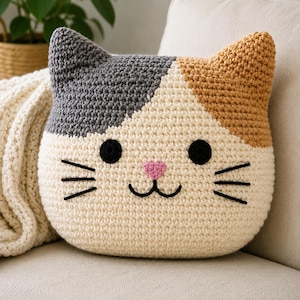 Puede incluir: Un cojín de ganchillo con forma de gato tricolor que presenta una cara de color crema, una mancha gris en la oreja izquierda y una mancha bronceada en la derecha. El cojín tiene dos ojos negros redondos, una nariz rosa y bigotes bordados.