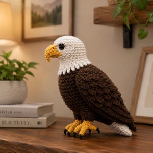 Puede incluir: Una figura de águila calva tejida a ganchillo con cabeza blanca, cuerpo marrón oscuro, pico amarillo curvado y garras amarillas con uñas negras. El ave de peluche está hecha con puntadas visibles y se encuentra en posición de perfil.