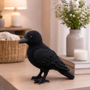 May include: A black crochet crow figurine featuring a textured yarn surface, a pointed beak, and small dark eyes. The bird is crafted with detailed stitching to mimic feathers on the wings and body, standing on two small, dark, multi-toed feet.
