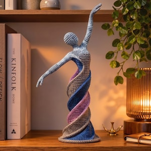 May include: A crochet sculpture of a dancing figure with one arm raised, featuring a spiraling base in shades of light blue, grey, dark blue, and soft purple. The figure has a rounded head and slender arms, with a textured yarn surface throughout.
