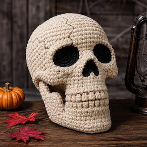 Puede incluir: Una calavera de ganchillo beige con cuencas oculares negras profundas y una cavidad nasal negra. La calavera presenta costuras detalladas que imitan la estructura del hueso humano, incluyendo la mandíbula y los dientes, hecha de hilo con un patrón texturizado.