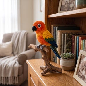 May include: A crocheted sun conure parrot figurine featuring a bright orange and yellow head, a black beak, and wings with yellow, green, and blue yarn. The bird is perched on a natural wooden branch stand with gray crocheted feet.