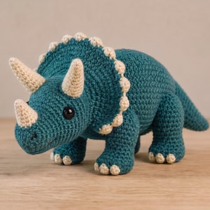 May include: A teal triceratops dinosaur toy made of crocheted yarn. The creature features three cream-coloured horns, a large frill with a scalloped cream edge, and small cream-coloured claws on each of its four feet. It has two black eyes.