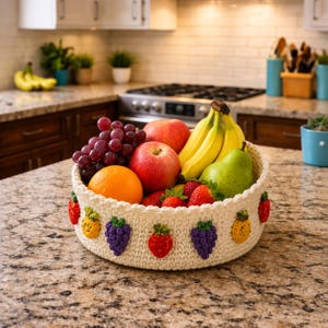 Può includere: Un cesto rotondo all'uncinetto color crema con piccole applicazioni di frutta colorate e testurizzate, tra cui fragole, uva e ananas, riempito con mele rosse fresche, banane, una pera verde, uva e un'arancia.